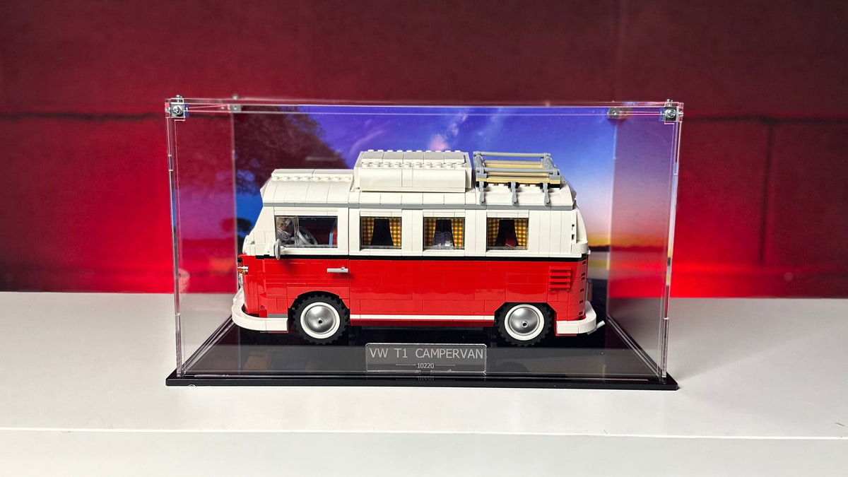 A red and white toy model of a classic Volkswagen Type II camper van displayed inside a clear acrylic shadowbox case on a black base, set on a white shelf against a red brick wall background.
