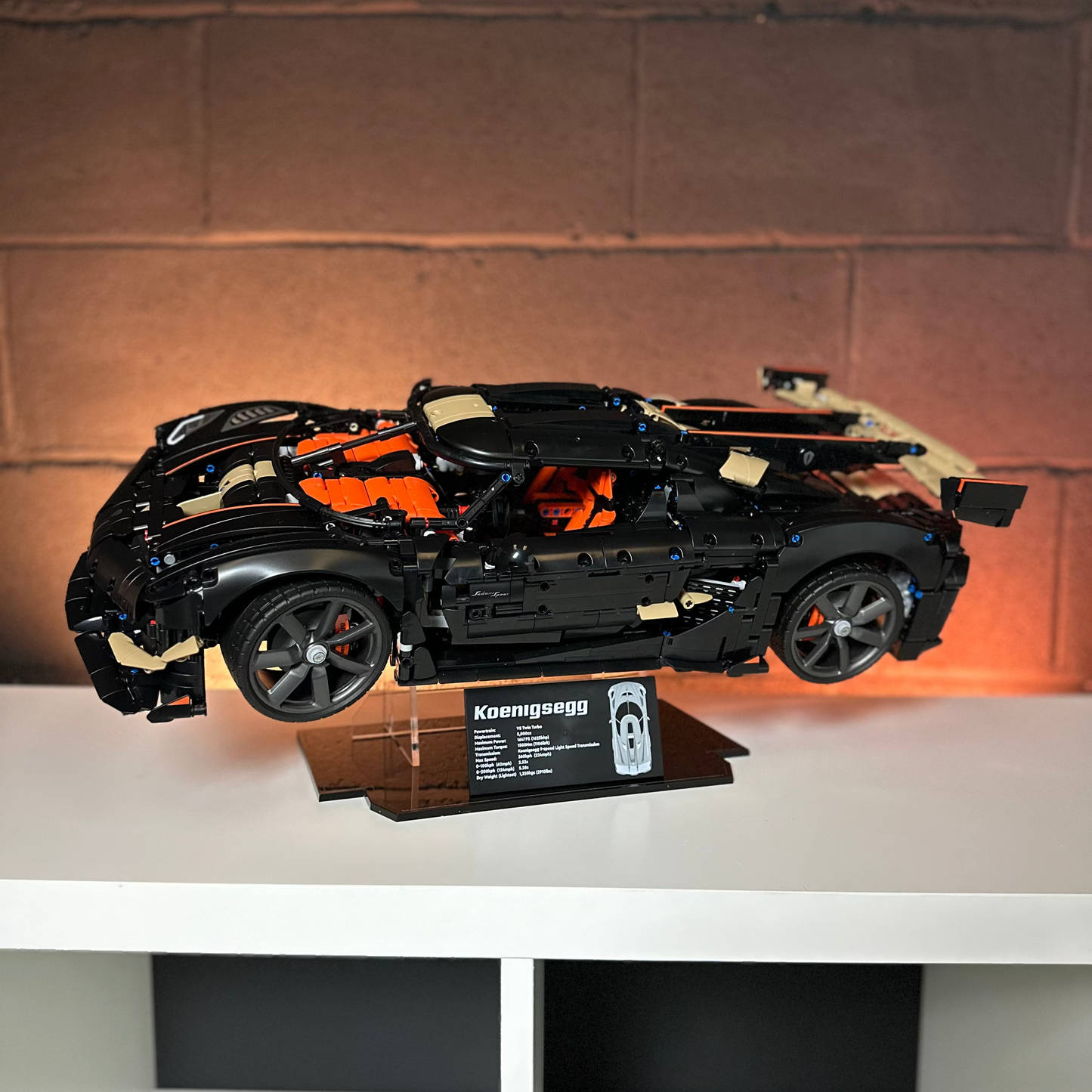 Koenigsegg LEGO model sports car displayed on a clear acrylic stand with a nameplate, sitting on a white shelf against a brick wall background.